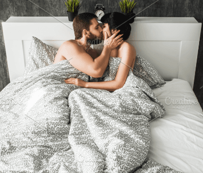 A couple kissing while lying together in bed under a patterned duvet